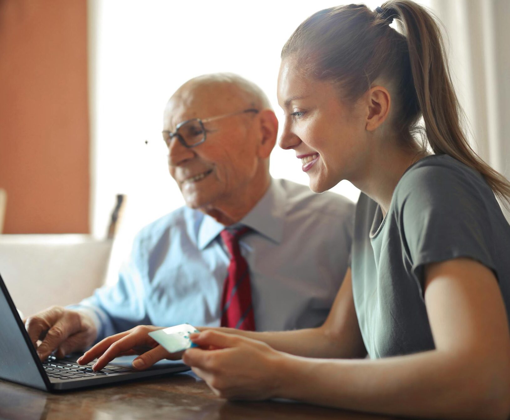 Young woman in casual clothes helping senior man in formal shirt with paying credit card in Internet using laptop while sitting at table